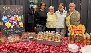4 women standing behind a table with signs, juice, and muffins